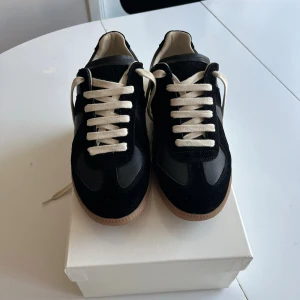 Maison Margiela Gats Black Gum - Stilrena svarta sneakers från Maison Margiela med vita kontrastsnören och beige sula. Colorway: Black Gum. Skorna har en klassisk låg silhuett och är tillverkade i en mix av mocka och läder för en lyxig känsla. Storlek 42. Hör av dig vid minsta fundering!