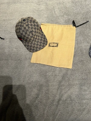 Gucci beige GG-mönstrad keps - Säljer en beige keps från Gucci med klassiskt GG-monogram över hela kepsen. Kepsen har böjd skärm och detaljer i rött och grönt på sidorna. Tillverkad i canvasmaterial och levereras med original dustbag. Perfekt för dig som gillar exklusiva accessoarer.