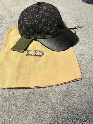 Svart keps från Gucci med GG-mönster - Snygg svart keps från Gucci med klassiskt GG-monogram i grått över hela kepsen. Skärmen är i svart läder och kepsen har en justerbar rem bak. Kommer med originalpåse och tagg. Perfekt för dig som vill ha en exklusiv och trendig accessoar.