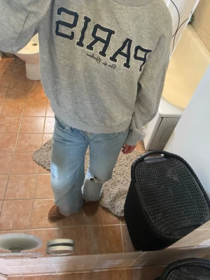 Grå sweatshirt med Paris-tryck - Säljer en grå sweatshirt med stort Paris-tryck i mörkblått på bröstet och text under. Tröjan har rund hals, långa ärmar och ribbade muddar vid ärmslut och nederkant. Perfekt för dig som gillar en chill och trendig look. Materialet känns mjukt och bekvämt. Lite defekt i brodyren.