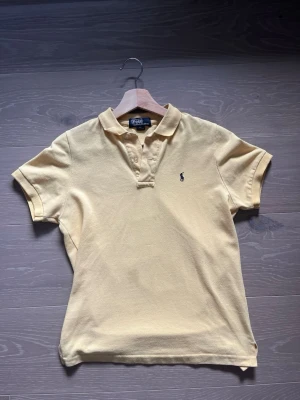 Gul pikétröja från Ralph Lauren - Klassisk gul pikétröja från Ralph Lauren med korta ärmar och krage. Tröjan har knappar vid halsen och den ikoniska broderade loggan på bröstet. Tillverkad i 100% bomull för en skön och luftig känsla.