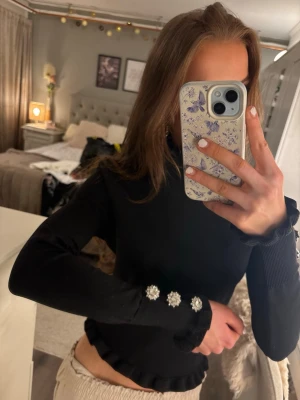 Svart långärmad topp med pärlknappar - Trendig svart långärmad topp med volangkant vid hals och midja. Ärmarna har dekorativa pärlknappar som ger en extra detalj. Toppen har en slim passform och är perfekt för dig som gillar stilrena plagg med det lilla extra.