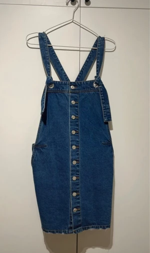 Blå jeansklänning med knappar - Cool blå jeansklänning med axelband och knappar hela vägen framtill. Klänningen har fickor både fram och bak samt en rak passform. Perfekt för att styla med t-shirt eller tröja under. Snygg och enkel att bära.