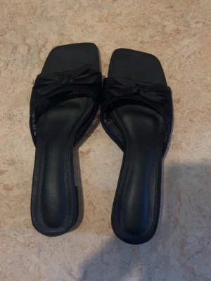 Svarta slip-in tofflor med rosett - Svarta slip-in tofflor med fyrkantig tå och dekorativ rosett på ovansidan. Tillverkade i syntetmaterial med ljus sula och låg klack. Perfekta för en avslappnad och trendig look.