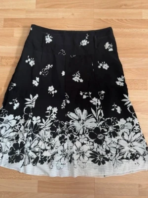 Svart blommig kjol från Lisa David - Svart kjol från Lisa David med vita blommor och diskret mönster längst ner. Kjolen har en klassisk A-linjeform och är knälång. Perfekt för dig som gillar stilrena och snygga plagg med blommigt mönster. Tygg bälte saknas. Man kan använda utan elle kombinera med eget vit bältet.