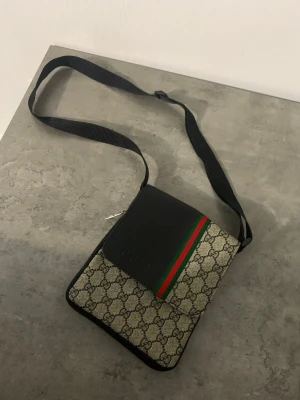 Axelväska  - Snygg axelväska med klassiskt GG-mönster i beige och svart, samt detaljer i svart skinn. Väskan har gröna och röda ränder på framsidan och en praktisk dragkedja på baksidan. Perfekt storlek för mobil och småsaker.