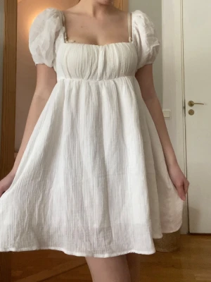 Vit kort klänning med puffärmar - Supersöt vit kort klänning med puffärmar och fyrkantig urringning. Klänningen har en luftig och flowy passform, tunna axelband och är gjord i ett lätt, strukturerat bomullstyg. Perfekt för varma dagar och ger en romantisk vibe.