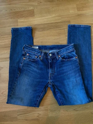 Blå Levi's straight fit jeans w28 L32 - Säljer ett par klassiska blå Levi's jeans med straight fit och fem fickor. Jeansen har orangea sömmar och en brun läderpatch bak i midjan. Materialet är slitstark denim och passformen är tidlös och enkel. Storlek w38 L32