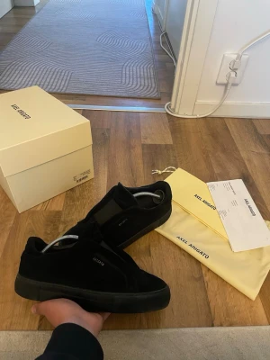 Svarta sneakers från Axel Arigato - Snygga svarta sneakers från Axel Arigato med diskret logga på sidan. Skorna har klassisk snörning och är tillverkade i mocka med en svart sula. Perfekta för dig som gillar stilrena och minimalistiska skor. Mycket finnt skick obs box medföljer ej 