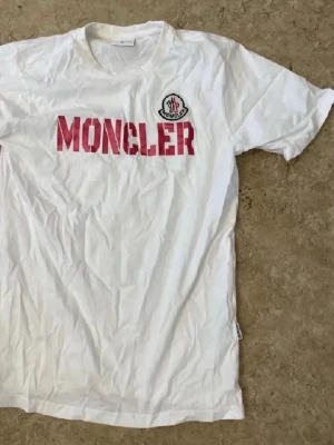 Vit Moncler t-shirt med röd logga - Vit t-shirt från Moncler med röd MONCLER-tryck på bröstet och klassisk logotyp på vänster sida. Rund halsringning och korta ärmar. Perfekt för dig som gillar stilrena och exklusiva plagg.