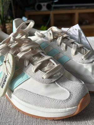 Adidas sneakers med mintblå ränder 38 - Snygga Adidas sneakers i vitt och ljusgrått med mintblå ränder på sidan. Ovandelen är i en mix av mocka och textil, och sulan är i gummi med klassiskt mönster. Skorna har vita snören och retrovibbar som passar perfekt till streetwear. Storlek 38