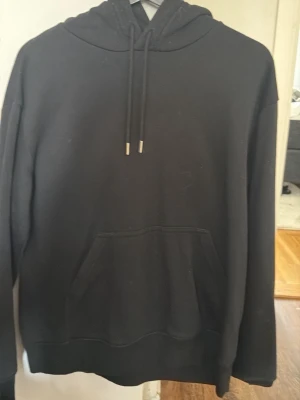 Svart hoodie från H&M - En klassisk svart hoodie från H&M med huva och dragsnören. Tröjan har en stor magficka framtill och ribbade muddar vid ärmslut och nederkant. Perfekt för en avslappnad och cool look. Materialet är mjukt och bekvämt, troligen bomullsblandning.