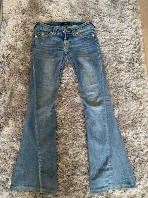 Blå bootcut jeans från True Religion - Jensen är uppsydda!! Innerbenslängden är 75cm.                                                           Säljer ett par blå bootcut jeans från True Religion med klassiska kontrastsömmar och guldfärgade knappar. Jeansen har snygga fickdetaljer bak och är tillverkade i denim med lätt tvättad look. Perfekt för dig som gillar en avslappnad och trendig stil.