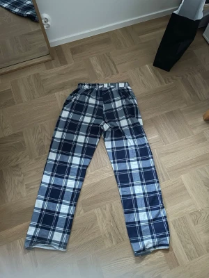 Blårutiga pyjamas byxor - Ett par sköna pyjamas byxor med blåvitt rutmönster och resår i midjan. Perfekta för att chilla hemma i. Byxorna har raka ben och är tillverkade i ett mjukt tyg som känns bekvämt mot huden.