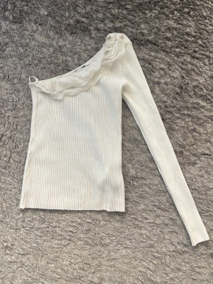 Vit oneshoulder topp med volang - Snygg ribbad vit oneshoulder topp med lång ärm och volangdetalj längs halsen. Materialet är mjukt och stretchigt, vilket gör den bekväm att bära. Den är väldigt stretchig så att den kan fungera både som xs, S och även m i vissa fall, har bara haft på mig den en gång 