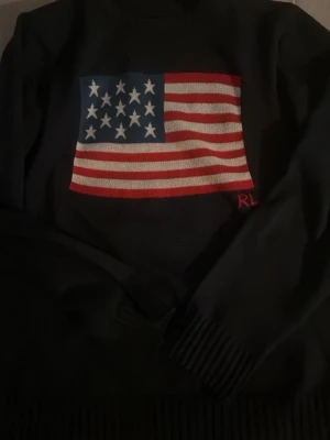 Mörkblå stickad tröja Ralph Lauren - Stickad mörkblå tröja från Ralph Lauren med stor amerikansk flagga på bröstet och RL-broderi i rött. Tröjan har långa ärmar och ribbade muddar vid ärmslut och nederkant. Perfekt för dig som gillar klassisk och tidlös stil.