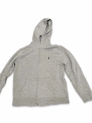 Grå zip-hoodie från Polo Ralph Lauren - Säljer en grå hoodie med dragkedja från Polo Ralph Lauren. Hoodien har en huva, två stora fickor fram och den klassiska broderade loggan på bröstet. Materialet är mjukt och bekvämt, perfekt för chill dagar. Enkel och stilren design som passar till det mesta.