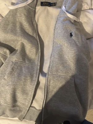Grå hoodie från Polo Ralph Lauren - Snygg grå hoodie från Polo Ralph Lauren med dragkedja, vita snören i huvan och klassisk broderad logga på bröstet. Tillverkad i mjuk bomullsblandning och har två stora fickor framtill. Perfekt för en avslappnad och stilren look. OBS den har ingen kedja. De går att lösa vid någon affär. 