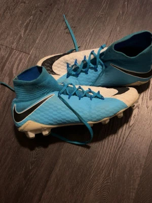 Nike blå-vita fotbollsskor med strumpa - Säljer ett par Nike fotbollsskor med hög strumpa och snörning. Skorna är blå och vita med svarta Nike-loggor på sidorna. De har plastdobbar och är tillverkade i syntetmaterial med textur för bättre bollkontroll. Perfekta för spel på gräs.Storlek 43