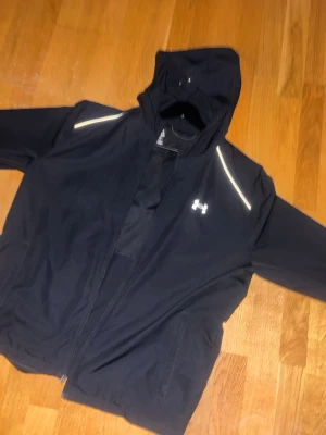 Svart vindjacka från Under Armour - Snygg svart vindjacka från Under Armour med huva och dragkedja. Jackan har reflekterande detaljer på axlarna och logga på bröstet. Tillverkad i lätt och tåligt material, perfekt för blåsiga dagar. Två sidofickor och sportig passform.
