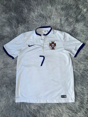 Portugal Ronaldo matchtröja Nike 2014 authentic  - Vit Portugal landslagströja från Nike med blå detaljer, nummer 7 och Ronaldo på ryggen. Tröjan har korta ärmar, knappslå vid halsen och landslagets emblem på bröstet. Materialet är lätt och andas, perfekt för fotboll och sport.