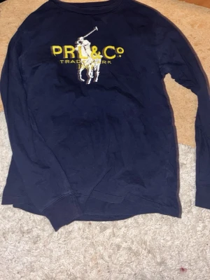 Mörkblå långärmad tröja Polo Ralph Lauren - Mörkblå långärmad tröja från Polo Ralph Lauren med stort broderat motiv av en polospelare och texten PRL & Co i gult på bröstet. Tröjan har rund hals och är tillverkad i mjukt material som passar perfekt för en avslappnad look.