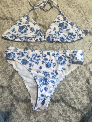 Blåvit blommig bikini med knyt - Snygg bikini med blommigt mönster i blått och vitt. Överdelen har triangelformade kupor och knytband i nacken. Underdelen är klassisk med hög skärning. Perfekt för strandhäng och poolparty!