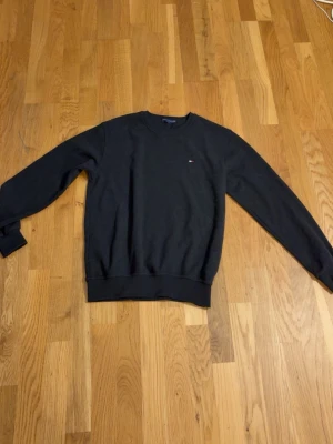 Svart sweatshirt från Tommy Hilfiger - En stilren svart sweatshirt från Tommy Hilfiger med klassisk rund hals och diskret logobrodyr på bröstet. Tröjan har ribbade muddar vid ärmslut och nederkant, perfekt för en avslappnad look. Materialet är mjukt och bekvämt.