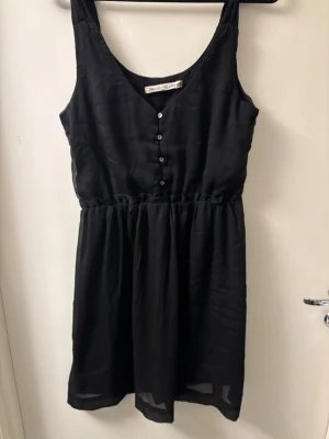 Svart kortklänning med knappar - Svart kortklänning från H&M med v-ringad front och dekorativa knappar. Klänningen är ärmlös och har en lätt rynkad midja som ger en snygg siluett. Materialet är tunt och luftigt, perfekt för varma dagar. Enkel och stilren design som är lätt att matcha.