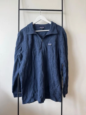 Blå långärmad tröja från Patagonia - En blå långärmad tröja från Patagonia med halv dragkedja och krage. Tröjan har ett diskret logomärke på bröstet och är tillverkad i ett mjukt, strukturerat material. Perfekt för lager-på-lager och chill stil.