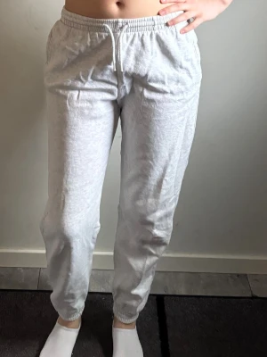 Ljusgrå mjukisbyxor med dragsko - Ljusgrå mjukisbyxor med elastisk midja och vit dragsko. Byxorna har raka ben med muddar vid anklarna och sidofickor. Perfekta för chill och avslappnade dagar, tillverkade i mjukt bomullsmaterial.