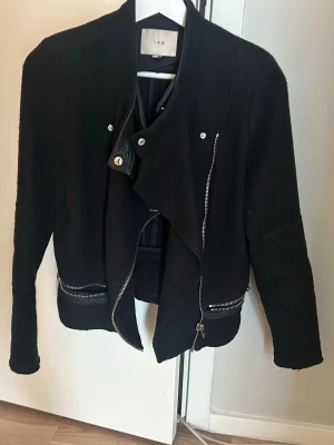 Svart skinnjacka från IRO - Snygg svart ulljacka från IRO med svarta skinndetaljer, asymmetrisk dragkedja, dekorativa silverfärgade dragkedjor och knappar. Jackan har e bikerstil och detaljer vid kragen. Perfekt som vårjacka! Köpt i Paris, storlek 40 FR mrn jag skulle säga en M eller vanlig storlek 38. 