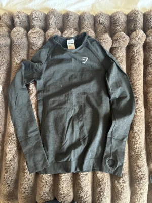 Mörkgrå långärmad tröja Gymshark - Mörkgrå långärmad tröja från Gymshark med diskret mönster och vitt logotryck på bröstet. Tröjan har rund halsringning och är tillverkad i ett stretchigt och ventilerande material som passar perfekt för träning och sport.