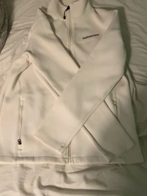 Peak Performance tröja - Säljer en stilren vit hoodie från Peak Performance. Hoodien har dragkedja framtill, två fickor med dragkedja och en liten logga på ryggen. Tillverkad i ett mjukt och stretchigt material som känns skönt mot huden. Perfekt för dig som gillar enkel och clean stil.
