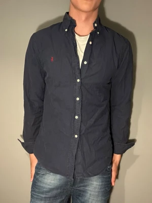 Mörkblå skjorta från Polo Ralph Lauren - Säljer en mörkblå skjorta från Polo Ralph Lauren med klassisk knappkrage och vita knappar. Skjortan har lång ärm och den ikoniska röda logon broderad på bröstet. Perfekt för dig som gillar stilrena plagg med snygga detaljer.