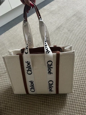 Chloé vit canvas handväska - Snygg vit canvas handväska från Chloé med bruna detaljer och breda vita handtag med Chloé-logga. Väskan har en boxig form och är perfekt för dig som gillar stilrena accessoarer med designer-touch.
