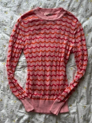 Rosa stickad tröja med zigzag-mönster - Säljer en tunnstickad tröja i rosa, rött och orange med vågigt zigzag-mönster. Tröjan har rund halsringning och långa ärmar 