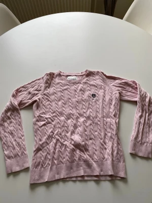 Ljusrosa kabelstickad tröja Bondelid - Ljusrosa kabelstickad tröja från Bondelid med rund hals och långa ärmar. Tröjan har ribbade muddar och ett diskret Bondelid -emblem på bröstet. Perfekt för att addera lite färg till din garderob. Materialet känns mjukt och bekvämt.