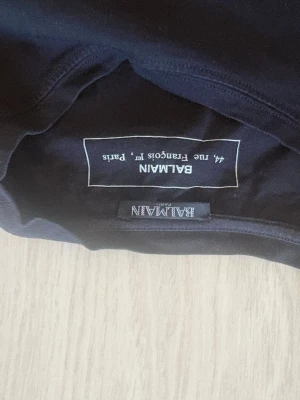 Svart Balmain t-shirt med guldtryck - Snygg svart t-shirt från Balmain med stort guldigt logotryck på bröstet. Klassisk rund halsringning och korta ärmar. Perfekt för dig som vill ha en exklusiv och trendig look. Materialet är mjukt och känns skönt mot huden.