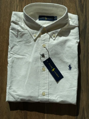 Vit skjorta från Ralph Lauren - Klassisk vit skjorta från Ralph Lauren med button-down krage och broderad marinblå logga på bröstet. Skjortan har långa ärmar, knappar framtill och är tillverkad i mjuk bomull. Perfekt för en clean och stilren look.