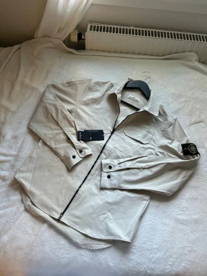Beige overshirt från Stone Island - Stilren beige overshirt från Stone Island med svart dragkedja, bröstficka och klassisk logopatch på vänster ärm. Tillverkad i ett lätt, slätt material som ger en modern look. Perfekt för lager-på-lager och streetwear-vibe.