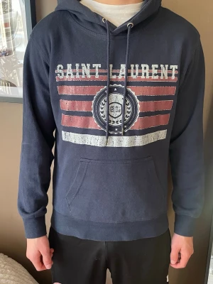 Mörkblå Saint Laurent hoodie - Snygg mörkblå hoodie från Saint Laurent med tryck i vitt och rött framtill. Klassisk känguruficka och justerbar huva med dragsko. Tillverkad i mjukt bomullsmaterial som känns skönt mot huden. Perfekt för en avslappnad och trendig look.