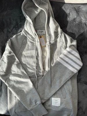 Thom browne zip up hoodie (Gray/1) - Snygg grå hoodie från Thom Browne med dragkedja framtill, justerbar huva och vita signaturränder på ena ärmen. Tillverkad i mjuk bomullsmix och har kängurufickor samt en liten logolapp nedtill. Perfekt för en avslappnad och trendig look.  Låt mig veta om du vill ha fler bilder. Skriv om man ör intresserad ;) KVITTO FINNS!