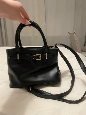 Svart liten handväska med axelrem - Snygg svart handväska i slätt konstläder med både handtag och justerbar axelrem. Väskan har detaljer i guld, samt en liten innerficka. Köpte i Portugal!