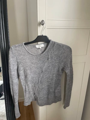 Grå kofta med knappar från Gina Tricot - Mjuk grå kofta från Gina Tricot i ullblandning. Koftan har rund halsringning och dekorativa knappar som går snett över framsidan. Långärmad och perfekt för lager-på-lager. Enkel och stilren design som är lätt att matcha.
