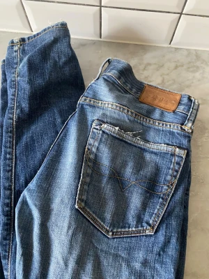 Ralph Lauren Jeans  - Säljer ett par riktigt snygga jeans från Polo Ralph Lauren i en perfekt vintage-tvätt! 👖 Dessa har den där klassiska, rejäla denimkänslan med snygga slitna detaljer vid fickorna och bensluten för en medvetet "lived-in" look. ✨ Detaljer: • Märke: Polo Ralph Lauren 🏇 • Storlek: W30 / L30 📏 • Färg: Mellanblå med naturlig patina och vintage-tvätt. • Modell: Klassisk passform med knappar i gylfen (button fly) och ikonisk läderpatch baktill. 🏷️ 