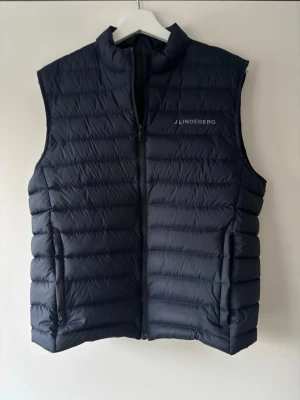 Mörkblå dunväst från J.Lindeberg - Stilren Mörkblå dunväst från J.Lindeberg med diskret logga på bröstet. Västen har en hög krage, dragkedja framtill och quiltad design som ger en modern look. Perfekt nu under våren eller till sommaren! Skriv om du har funderingar, pris kan diskuteras. Storlek M men kan absolut bäras som storlek S.
