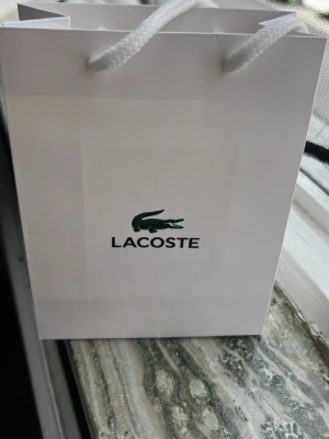 Silverfärgad klocka från Lacoste - Stilren silverfärgad klocka från Lacoste med klassiskt metallarmband och minimalistisk design. Levereras med originalförpackning och en vit tygpåse med Lacoste-loggan. Perfekt för dig som gillar snygga accessoarer med ikonisk krokodil.