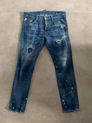 Blå distressed jeans från Dsquared2 - Säljer ett par blå jeans från Dsquared2 med slitna detaljer, hål och vita färgstänk. Jeansen har fem fickor, smal passform och coolt Dsquared2-märke på bakfickan. Perfekt för dig som gillar en edgy streetstyle-look.