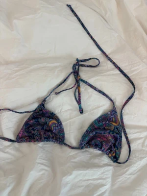 Färgglad bikiniöverdel med paisleymönster - Säljer en snygg bikiniöverdel från Primark med färgstarkt paisleymönster i blått, lila, grönt och gult. Triangelmodell med knytband i nacken och ryggen. Perfekt för strandhäng och poolparty. Materialet är polyester och syntet.